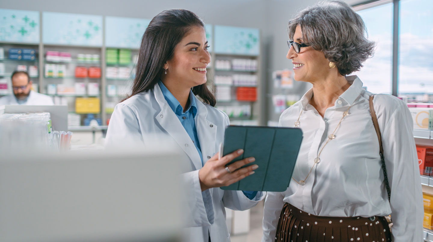 Pharmacienne en blouse blanche consultant un ouvrage de référence phytothérapie sur comptoir de pharmacie moderne, face à une patiente tenant une boîte de complément végétal, lumière naturelle douce